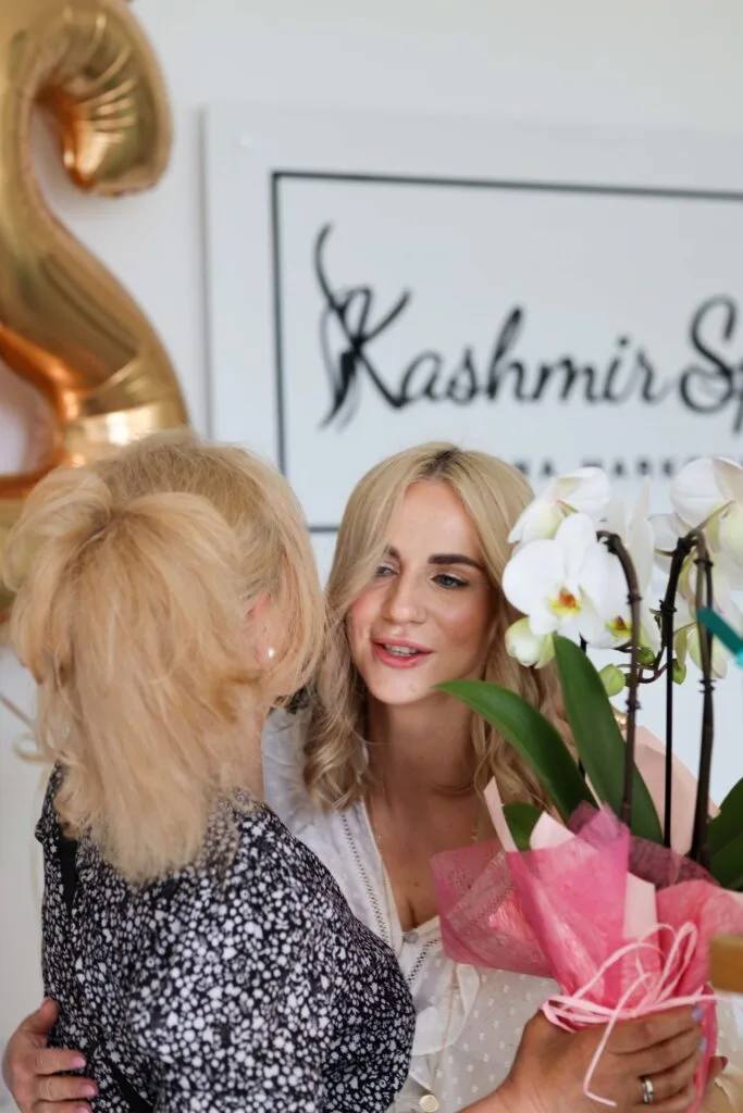 Dwie kobiety rozmawiające na 12 urodzinach Kashmir SPA, jedna trzyma bukiet kwiatów.