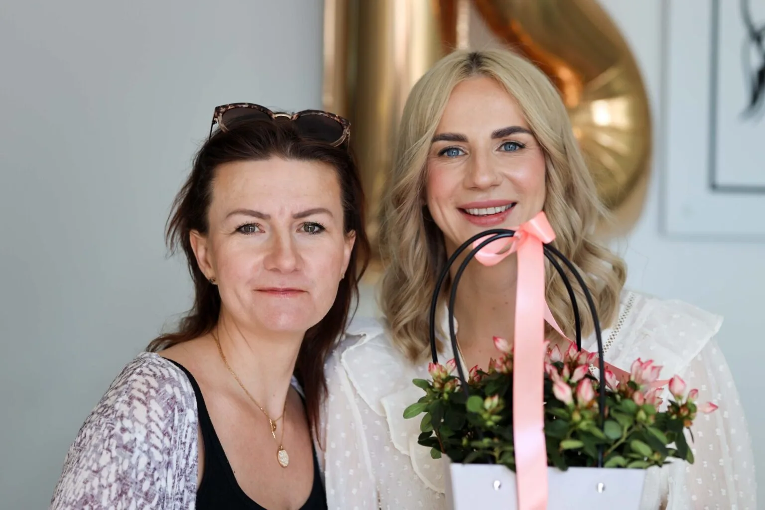 Dwie uśmiechnięte kobiety, jedna trzyma ozdobioną różową wstążką doniczkę z kwiatami otrzymaną na 12 urodziny salonu.