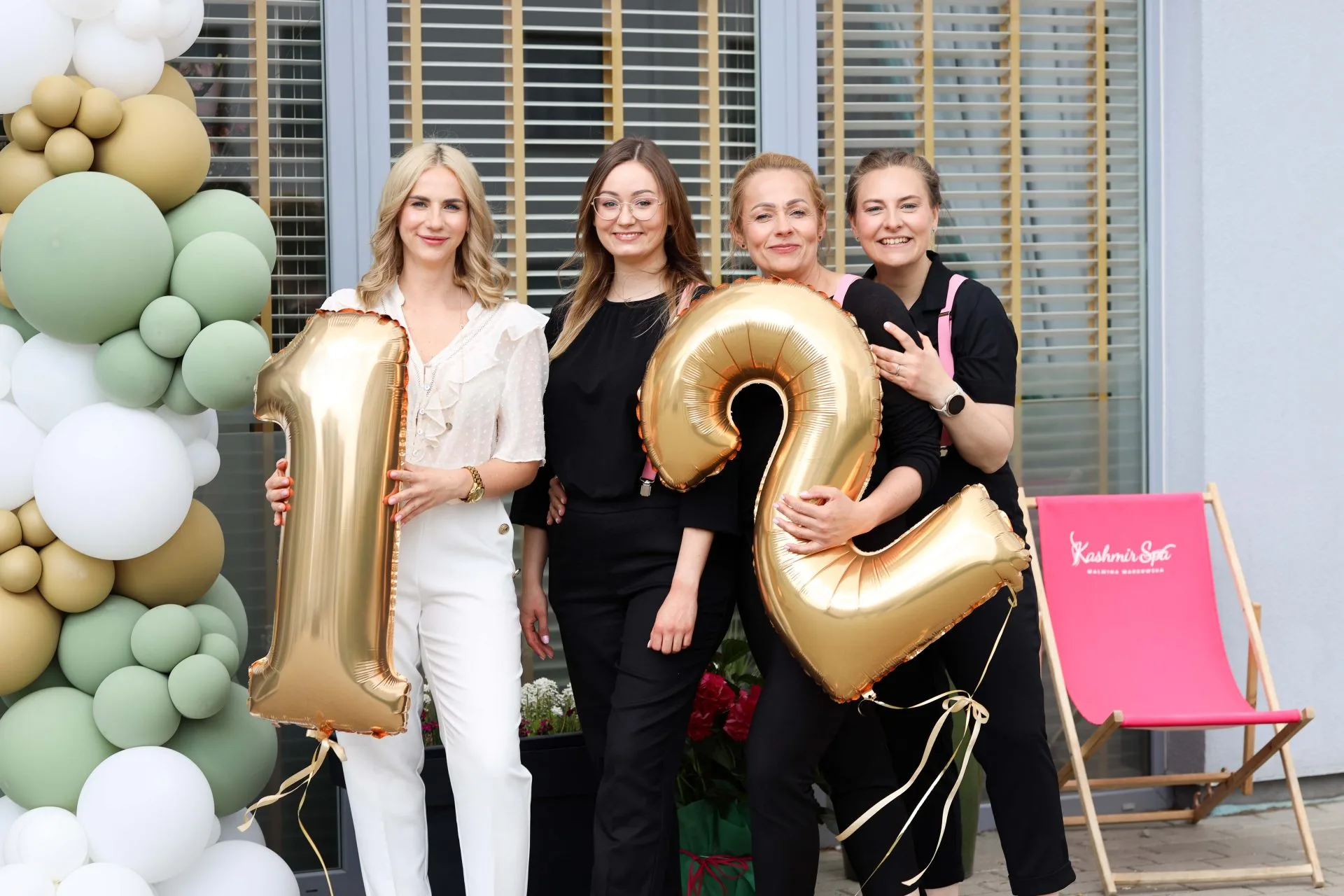 Cztery uśmiechnięte kobiety trzymają złote balony w kształcie cyfr 1 i 2, świętując 12 urodziny Kashmir SPA.