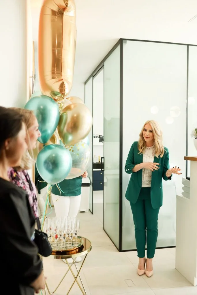 Właścicielka salonu w zielonym garniturze rozmawia z gośćmi przy stole z balonami i kieliszkami na 11 URODZINY salonu.