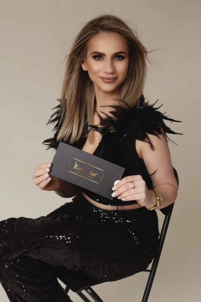 Kobieta trzymająca elegancką, czarną kopertę z voucherem na zabiegi w Kashmir Spa, ubrana w czarny strój z piórami.