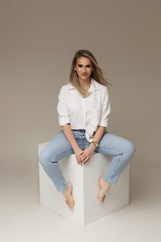 Właścicielka KASHMIR SPA Malwina Markowska, młoda kobieta w białej koszuli i dżinsach, siedzi na białym sześcianie w eleganckiej pozie